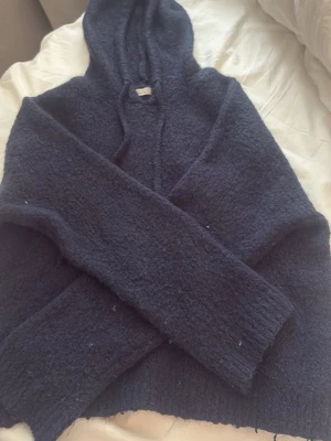 Mörkblå stickad hoodie - Mysig mörkblå stickad hoodie med luva och långa ärmar. Tröjan har en avslappnad passform och är tillverkad i ett mjukt, fluffigt material som känns riktigt skönt mot huden. Perfekt för dig som gillar en chill och enkel stil.