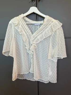 Vit blus med volanger från Zara - Söt vit blus från Zara i transparent, prickigt tyg med volangdetaljer längs knäppningen och kragen. Blusen har trekvartsärm och knäpps framtill med pärlemorknappar. Perfekt för en romantisk och trendig look.