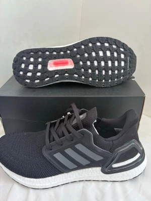 Adidas Ultraboost svarta sneakers - Svarta Adidas Ultraboost sneakers med stickad ovandel, vita Boost-mellansulor och svarta yttersulor. Skorna har de klassiska tre ränderna på sidan och en stabil hälkappa i plast. Perfekta för dig som gillar sportig och modern stil.