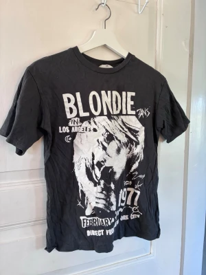 Svart Blondie t-shirt för flickor från H&M - Cool svart t-shirt för flickor från H&M med stort vitt Blondie-tryck framtill och text om en konsert i Los Angeles 1977. Klassisk rund hals och korta ärmar. Perfekt för dig som gillar bandtröjor och vill ha en avslappnad stil.