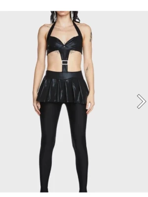 Svart wetlook klänning från Dolls kill stl XS - Säljer en klänning i wetlook-material med halterneck-top, dekorativt spänne framtill, kort plisserad kjol. Perfekt för raveparties eller festivaler där du vill sticka ut med en edgy och unik look.Leggins ingår ej.