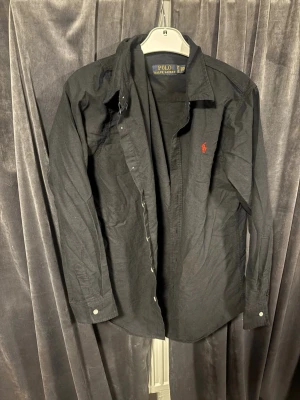 Svart skjorta från Polo Ralph Lauren - Klassisk svart skjorta från Polo Ralph Lauren med röd broderad logga på bröstet. Skjortan har lång ärm, knappar framtill och en tidlös krage. Tillverkad i mjuk bomull som känns skön mot huden.
