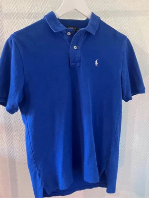 Blå pikétröja från Ralph Lauren - Klassisk blå pikétröja från Ralph Lauren med vit broderad logga på bröstet. Tröjan har korta ärmar, krage och knäppning med två knappar framtill. Bra skick
