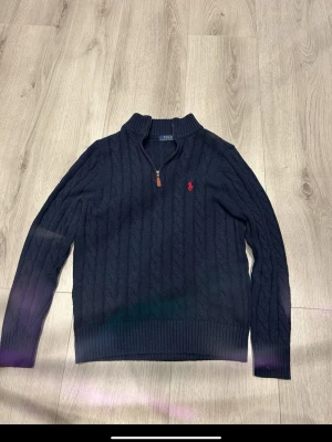 Mörkblå kabelstickad tröja Polo Ralph Lauren - Kabelstickad mörkblå tröja från Polo Ralph Lauren med halv dragkedja och klassisk röd logga broderad på bröstet. Tröjan har ribbade muddar och hög krage. Perfekt för dig som gillar stilrena och tidlösa plagg i mjukt material.