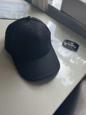 Svart keps från Fendi med logomönster - Snygg svart keps från Fendi med diskret ton-i-ton logomönster över hela kepsen. Klassisk böjd skärm och justerbar baksida. Tillverkad i ett slitstarkt tyg som ger en exklusiv känsla. Perfekt accessoar för en trendig streetwear-look.
