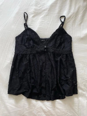Svart topp från Gina tricot - Snygg svart topp från gina tricot med tunna axelband och v-ringning. Linnet har två små knappar framtill och är tillverkat i ett lätt material med blommigt spetsmönster. Svinbra till varma sommarkvällar, bara aldrig kommit till användning❣️