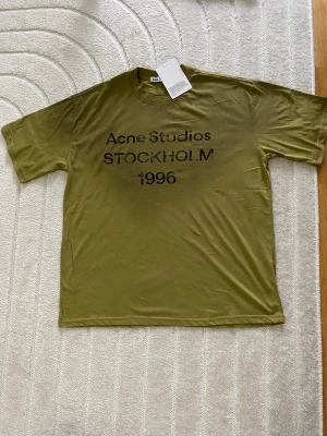 Olivgrön Acne Studios t-shirt - Olivgrön t-shirt från Acne Studios med svart text 'Acne Studios STOCKHOLM 1996' på bröstet. Klassisk passform med rund hals och korta ärmar. Tillverkad i mjuk bomull, perfekt för en clean och avslappnad stil.
