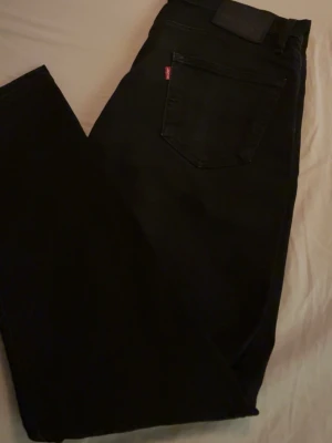 Svarta jeans från Levi's - Säljer ett par svarta jeans från Levi's med klassisk femficksdesign och rak passform. Jeansen har den ikoniska röda Levi's-lappen på bakfickan och är tillverkade i ett slitstarkt bomullsmaterial. Perfekta för en clean och stilren look.