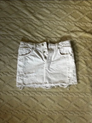 Vit jeanskjol - Kort vit jeanskjol med fem fickor, slitna detaljer och rå, fransig nederkant. Kjolen har klassisk knapp- och dragkedjestängning framtill och är gjord i denim. Perfekt för en avslappnad och trendig look.