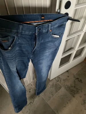 Blå jeans från BOSS med raka ben - Säljer ett par klassiska blå jeans från BOSS med raka ben och fem fickor. Jeansen har snygga slitningar och kontrastsömmar samt en läderpatch bak i midjan. W35/L32