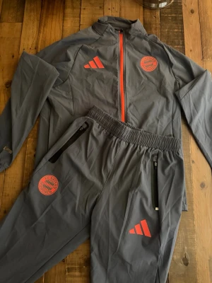 FC Bayern München träningsset - Säljer ett grått träningsset från Adidas x FC Bayern München. Setet består av en jacka och byxor med röda detaljer, klubbmärke och Adidas-logga. Jackan har hel dragkedja och fickor med dragkedja, byxorna har resår i midjan och dragkedjor vid fickorna. Perfekt för fotbollsträning.