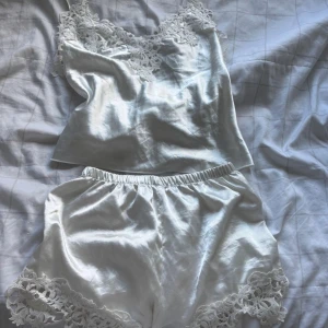 Vit satinpyjamas med spetsdetaljer - Snygg vit pyjamas i glansigt satinmaterial med spetsdetaljer vid linningen och bensluten. Setet består av ett linne med tunna axelband och matchande shorts med elastisk midja. Perfekt för mysiga kvällar hemma.
