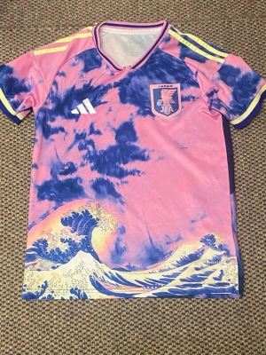 Japan fotbollströja rosa/blå Adidas - Unik Japan fotbollströja från Adidas i rosa och blått med vågmönster och gula detaljer. Tröjan har rund hals, korta ärmar och Japan-emblem på bröstet. Perfekt för dig som vill sticka ut på planen eller supporta landslaget.