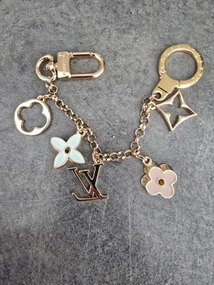Louis Vuitton guldfärgad väsksmycke - Snygg väsksmycke från Louis Vuitton i guldfärgad metall med flera ikoniska LV-hängen i vitt, ljusblått och rosa emalj. Kedjan har både karbinhake och ring för enkel fastsättning på väska eller nycklar. Perfekt accessoar för att lyfta din look.