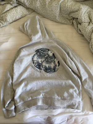 Grå hoodie med discokulor - Säljer en grå hoodie med tryck av två discokulor och texten 'DISCO CLUB' på ryggen. Hoodien har huva och ribbade muddar vid ärmslut och nederkant. Perfekt för dig som gillar coola och unika tryck. Materialet känns mjukt,bekvämt och är ganska tunnt.