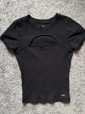 Svart ribbad topp med cut-out - Trendig svart ribbad topp med korta ärmar och en snygg cut-out detalj över bröstet. Figurnära passform och rund halsringning. Perfekt för dig som vill ha en edgy och stilren look.