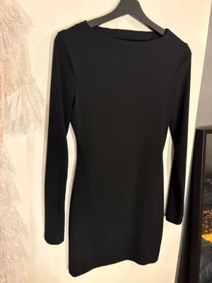 Svart tight långärmad klänning - Snygg svart klänning i stretchigt material med lång ärm och en djup, öppen rygg. Kort modell som sitter figurnära och har en enkel, stilren design. Perfekt för dig som gillar minimalistisk stil och vill ha något som sticker ut med öppen rygg.