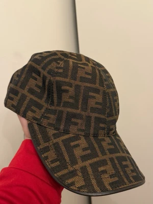 Brun Fendi keps - Säljer en brun keps från Fendi med det ikoniska FF-logomönstret i svart över hela kepsen. Kepsen har böjd skärm och justerbart band bak i brunt läder med metallspänne. Materialet är en mix av tyg och läderdetaljer. Perfekt för dig som gillar lyxiga accessoarer.