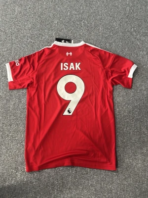 Liverpool matchtröja Isak #9 Adidas - Röd Liverpool matchtröja med vita detaljer, tryck 'ISAK' och nummer 9 på ryggen. Adidas-logga och Liverpool FC-märke på bröstet samt sponsortryck på framsidan. Tillverkad i lätt och ventilerande funktionsmaterial, perfekt för fotboll eller träning.