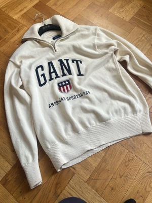 Vit halvzip tröja från GANT - Snygg vit stickad halvzip tröja från GANT med broderad logga och texten 'American Sportswear' framtill. Tröjan har hög krage, ribbade muddar och är tillverkad i bomull. Perfekt för en sportig och stilren look.