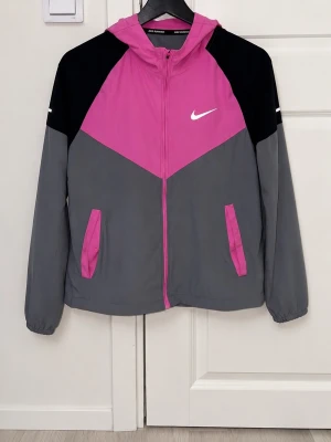 Ni och ke Miler rosa  - Snygg vindjacka från Nike i färgerna rosa, grå och svart. Jackan har huva, dragkedja framtill och två fickor med rosa detaljer. Ikonisk Nike-logga på bröstet. Perfekt för blåsiga dagar och sportiga outfits.
