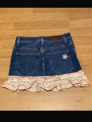 Jeanskjol med volang och blommor - Söt kort jeanskjol med volangkant i beige tyg med små blommor och rosa detaljer. Kjolen har broderade blommor fram och bak samt klassiska jeansfickor. Perfekt för dig som gillar en lekfull och trendig stil.