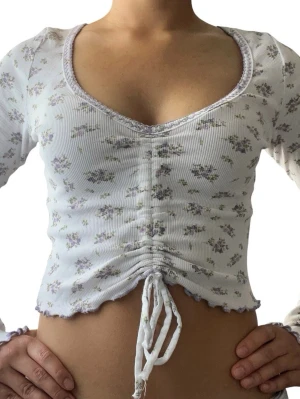 Blommig croppad topp med snörning - Vit croppad topp med små blommor i lila och grått. Toppen har lång ärm, ribbad struktur, v-ringning och snörning framtill som är justerbar.