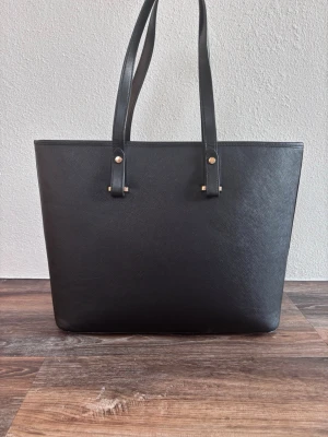 Svart shoppingväska i läderlook - Stilren svart shoppingväska med långsmala axelband och guldfärgade detaljer. Väskan har en klassisk rektangulär form och är tillverkad i ett material som liknar läder. Perfekt för att bära med sig allt man behöver under dagen.