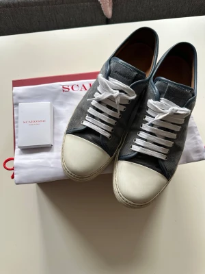 Scarosso gråa sneakers i mocka - Snygga sneakers från Scarosso i grå mocka med vita tåpartier och vita snören. Skorna har en klassisk låg profil och detaljer i läder på insidan. Perfekta för en stilren och avslappnad look.