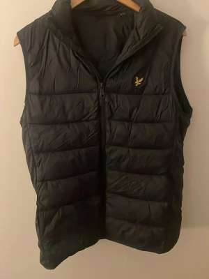 Svart dunväst från Lyle & Scott - Snygg svart dunväst från Lyle & Scott med gul broderad logga på bröstet. Västen har hög krage, dragkedja framtill och quiltad design. Perfekt för lager-på-lager och håller dig varm under kyliga dagar.
