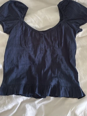 Mörkblå blus med puffärm och volang - Supersöt mörkblå blus med korta puffärmar, knappar framtill och volangkant nedtill. Blusen har en lätt rynkad byst och smockad rygg för extra komfort. Perfekt att styla med jeans eller kjol.