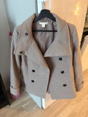 Beige dubbelknäppt rock från H&M - Stilren beige rock från H&M med dubbelknäppning och stora svarta knappar. Klassisk krage och figurnära passform. Perfekt för dig som gillar en enkel men snygg look. Tillverkad i ett mjukt tyg med slät yta.