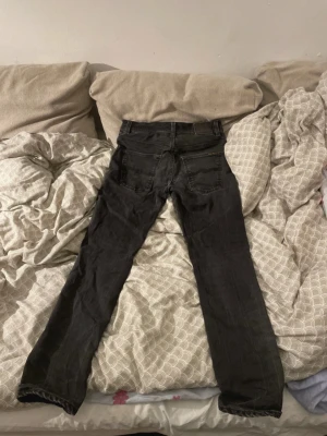 Svarta jeans från Nudie Jeans - Säljer ett par svarta jeans från Nudie Jeans med klassisk femficksdesign och diskret logga på bakfickan. Jeansen har raka ben och är tillverkade i denim med snygga slitningar. Perfekta för dig som gillar en stilren och tidlös look.