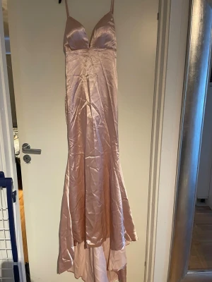Ljusrosa långklänning i satin - Elegant ljusrosa trumpet/sjöjungfru långklänning i glansigt satinmaterial med smala axelband och djup v-ringning. Klänningen har en figurnära passform upptill och faller snyggt längs kroppen. Perfekt för speciella tillfällen. Behöver bara ångas då den legat nedvikt. 