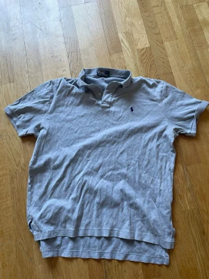 Grå pikétröja från Polo Ralph Lauren - Klassisk grå pikétröja från Polo Ralph Lauren med korta ärmar och krage. Tröjan har knappar vid halsen och den ikoniska broderade loggan på bröstet. Perfekt till jeans eller shorts för en avslappnad stil.