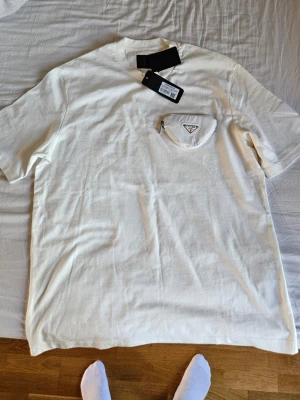 Vit t-shirt från Prada med ficka - Exklusiv vit t-shirt från Prada med en unik ficka med dragkedja och logga på bröstet. T-shirten har klassisk passform och korta ärmar. Perfekt för dig som vill sticka ut med en stilren och lyxig detalj.