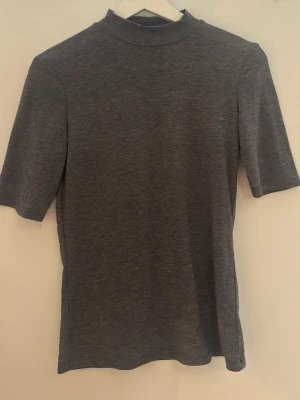 Grå polotopp från MNG - Snygg grå polotopp från MNG med korta ärmar och hög hals. Enkel och stilren design som passar perfekt till jeans eller kjol. Mjuk och bekväm i materialet, perfekt för lager-på-lager eller som den är.