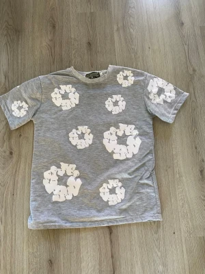 Grå t-shirt med vita blommor - Säljer en grå t-shirt med vita blommönster tryckta över hela plagget. T-shirten har rund hals och korta ärmar. Materialet känns mjukt och skönt, perfekt för en avslappnad stil. Passformen är normal och trycket ger en unik touch.