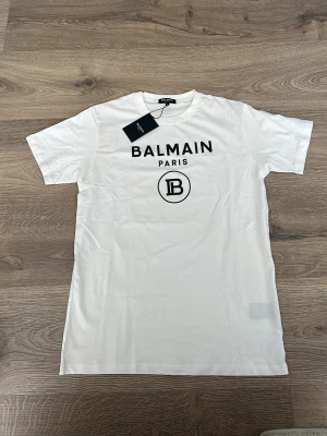 Vit Balmain Paris t-shirt - Snygg vit t-shirt från Balmain Paris med svart logotyptryck framtill. Klassisk rund halsringning och korta ärmar. Perfekt för dig som gillar stilrena och exklusiva plagg med tydlig branding.