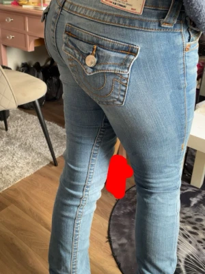 Lågmidjade botcut jeans från True Religion - Säljer ett par ljusblå jeans från True Religion med klassiska kontrastsömmar och ikonisk fickdesign bak. Jeansen har låg midja och är tillverkade i stretchigt denim för en tight passform. Perfekta för dig som gillar en trendig och avslappnad stil.