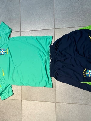 Brasilien Nike fotbollskit S - Snyggt Brasilien fotbollskit från Nike med ljusgrön t-shirt och mörkblå shorts. Båda plaggen har Brasilien-emblem och gula Nike-loggor. Shortsen har gröna detaljer och elastisk midja. Perfekt för träning eller match! Storlek S