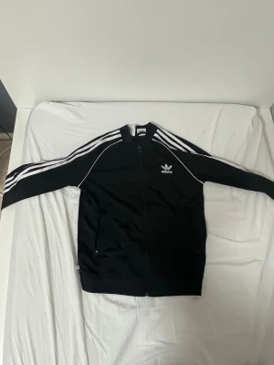 Svart Adidas SST track jacket - Klassisk svart Adidas track jacket med dragkedja, vita ränder längs ärmarna och Adidas logga på bröstet. Jackan har hög krage och är tillverkad i ett mjukt, sportigt material. Perfekt för träning eller streetwear.