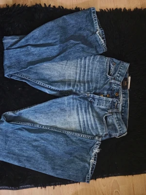 Blå vintage Lee jeans med raka ben - Säljer ett par klassiska blå Lee jeans i vintage-stil med raka ben och hög midja. Jeansen har knäppning med fyra kopparfärgade knappar och tydliga slitningar framtill för en cool look. Bakfickorna har Lee-logga i läder. Perfekta för en avslappnad streetwear-stil.