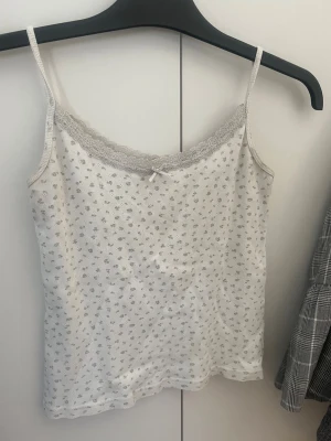Brandy Melville topp vit med blommor - Söt vit linnetopp med små blommor i grått och tunna axelband. Toppen har spetskant vid urringningen och en liten rosett framtill. Perfekt för en avslappnad och feminin look.