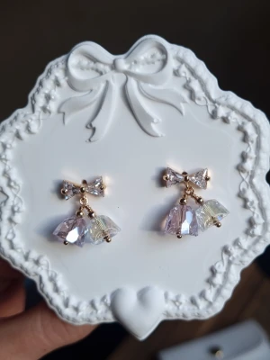 Aurora Drops - Dessa eleganta studs örhängen kombinerar en gnistrande rosett i kristall med hängande klockformade prismor. Kristallerna är slipade för att fånga varje ljusstråle och skapa en otrolig glans. Nickeltestat & allergivänligt Material: Rostfritt stål, Stift i 925 silver (stämplad)  Storlek Längd 2cm Bredd 1.9cm (rosett) -Levereras i ett smyckespåse som på bilden  - Inga prisförslag, tack                                       - Varan packas noggrant och säkert 