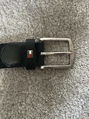 Svart Tommy Hilfiger bälte i skinn och textil - Snyggt svart bälte från Tommy Hilfiger med klassisk silverfärgad metallspänne. Bältet är gjort i en kombination av skinn och textil, med diskret logga på hällan. Perfekt för att lyfta din outfit med en stilren detalj.