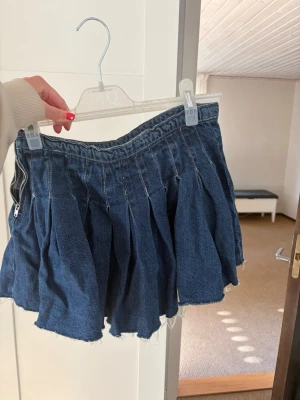 Blå plisserad jeanskjol  - Trendig blå jeanskjol med plisserad design och råa kanter. Kjolen har en kort modell och dragkedja i sidan. Perfekt för dig som gillar en avslappnad och cool stil. Slutsåld från Zara och köpt för två år sen.