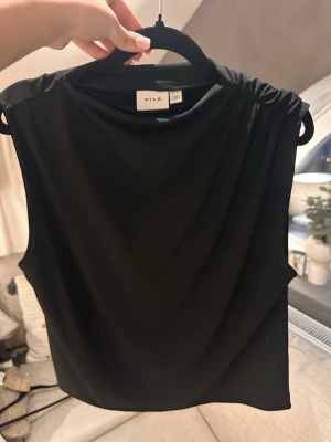 Svart ärmlös topp från Vila - Snygg svart topp från Vila med draperad axeldesign och rund halsringning. Toppen är ärmlös och är i ett mjukt tunt material. Ändast använd ett fåtal gånger.