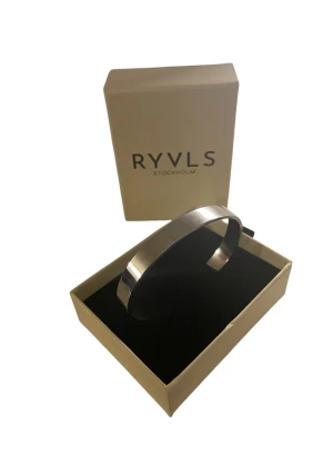 Silverfärgat armband från RYVLS - Stilrent och minimalistiskt armband i silverfärgad metall från RYVLS Stockholm. Armbandet har en slät, blank yta och är öppet i modellen vilket gör det enkelt att ta på och av. Perfekt accessoar för en modern look.