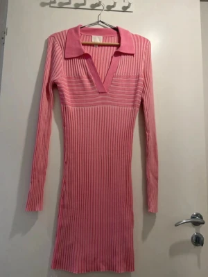 Rosa ribbad klänning från H&M - Rosa ribbad klänning från H&M med lång ärm och djup v-ringning. Klänningen har en klassisk krage och är figurnära med stretchigt material. Perfekt för dig som gillar en stilren och trendig look.